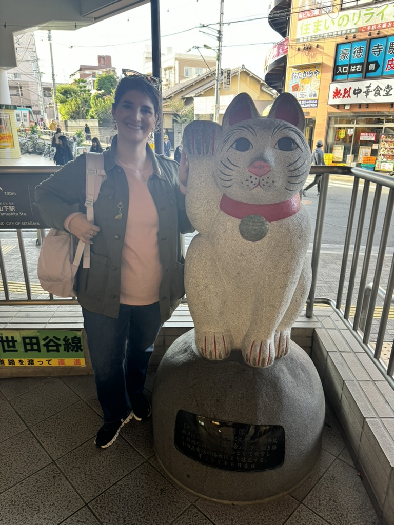 ashley posing with a giant maneki-neko (lucky cat) statue outside gotokuji station, where these iconic fortune-bringing cats originated